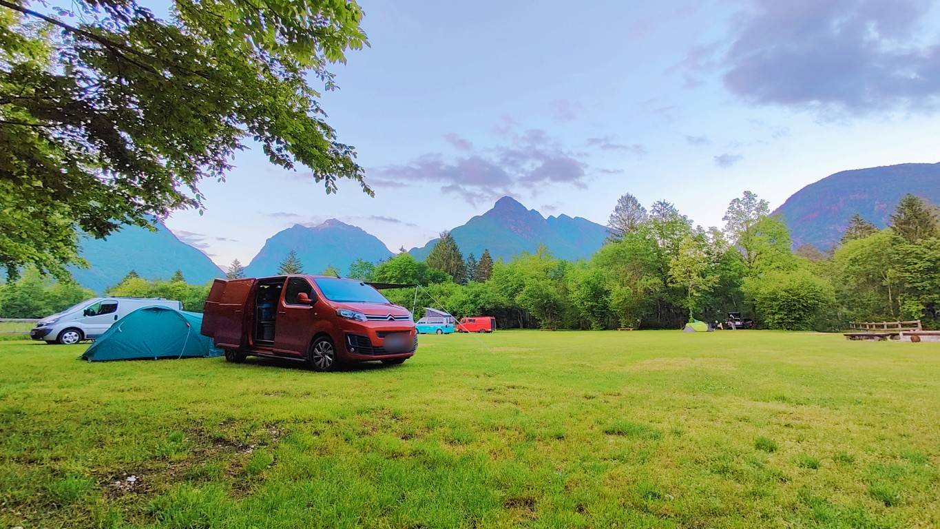 Umirjeno kampiranje v Kampu Vodenca, obdano z gozdom in gorami, z enostavnim kuriščem in avtodomi v naravi.