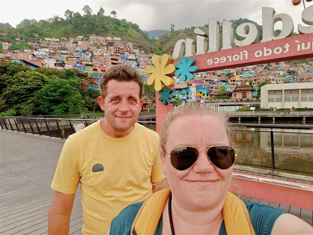Saša and Jure standing on a bridge in Medellín, Colombia, with colorful hillside houses in the background during their winter travels.